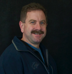 Brown haired man smiling with mustache in navy jacket with teal shirt